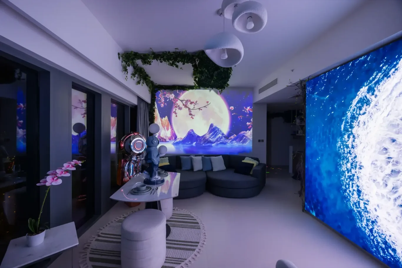 Modern living room with a large curved gray sofa, round ottomans, and a wall-wide screen displaying an ocean wave on the right and a moonlit landscape mural behind the seating area; green ivy decor overhead.