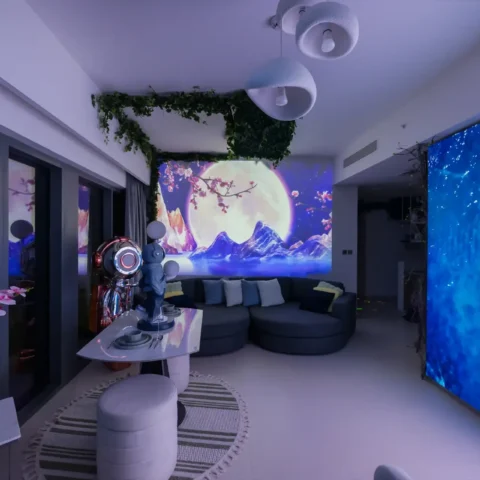 Modern living room with a large curved gray sofa, round ottomans, and a wall-wide screen displaying an ocean wave on the right and a moonlit landscape mural behind the seating area; green ivy decor overhead.