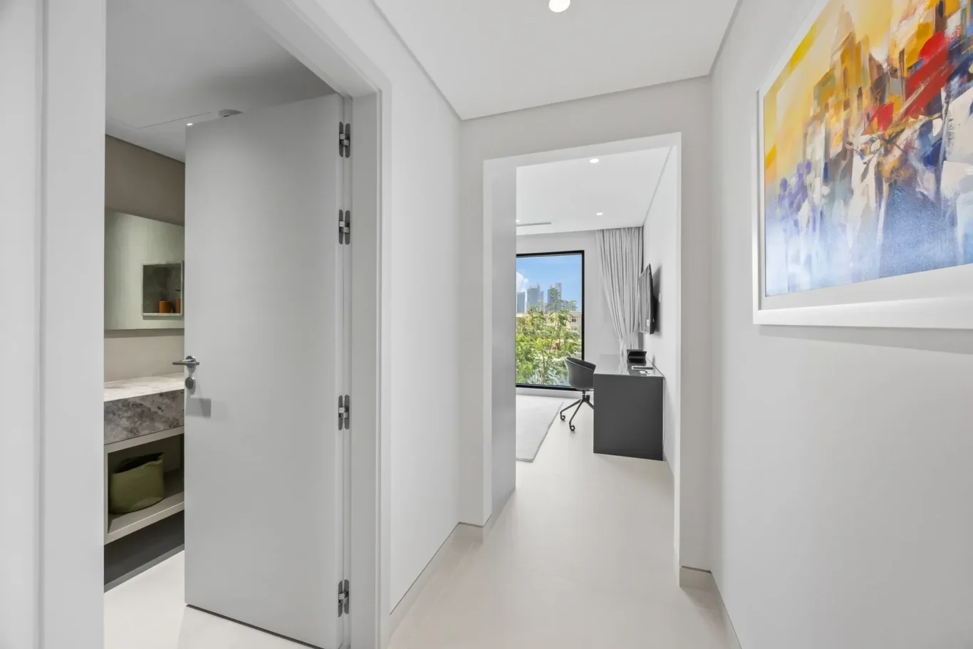 Bright hallway leading to a small office with desk and chair by a large window, colorful abstract painting on the right wall.