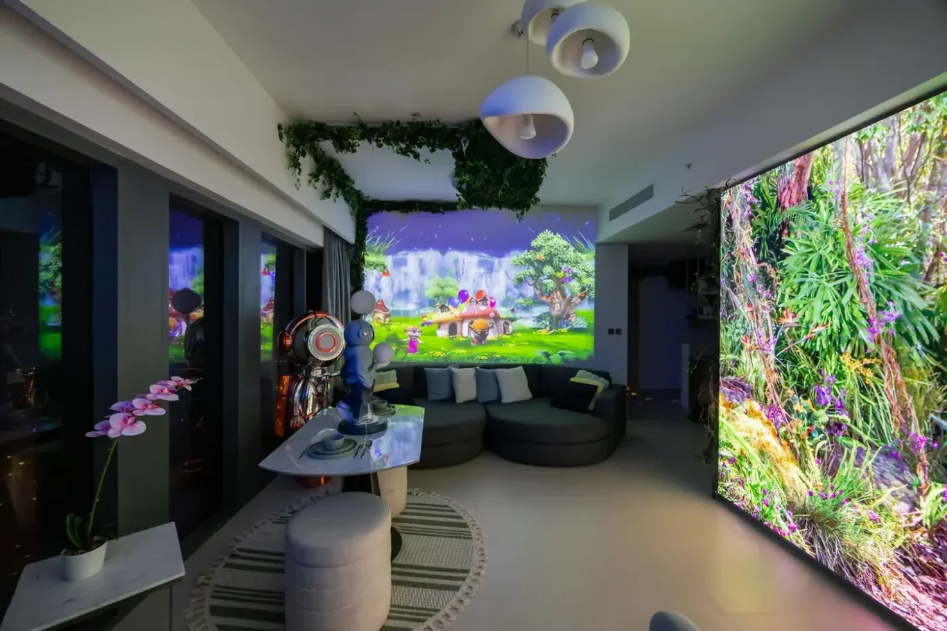 Modern living/entertainment room with a curved dark sofa, greenery overhead, and a colorful fantasy landscape projected on the back wall.