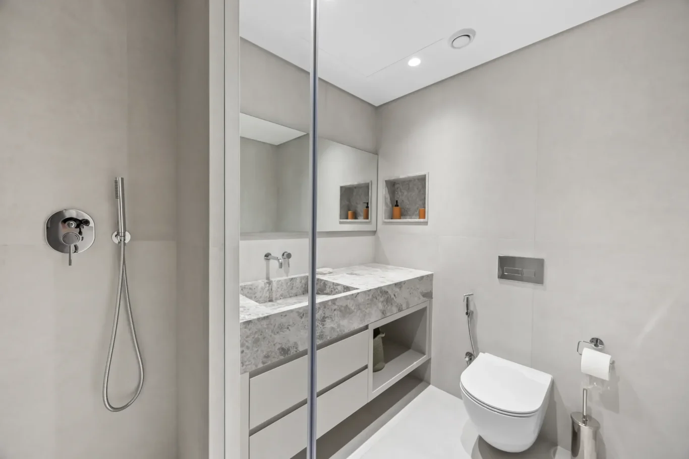 Modern gray bathroom with glass shower enclosure, marble vanity top, and wall-mounted fixtures; toilet on right with toilet roll holder nearby.