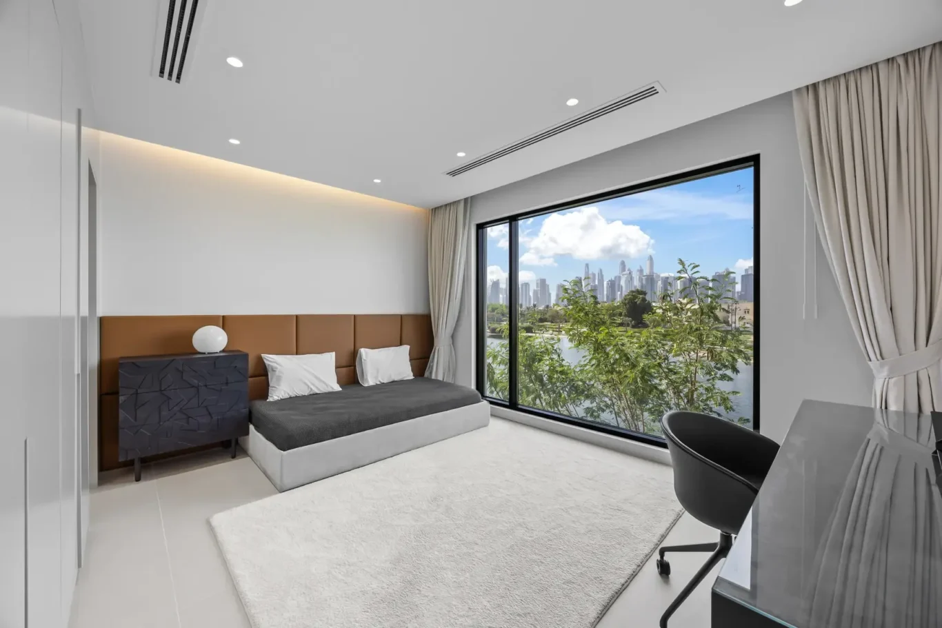 Modern bedroom with a large window overlooking a city skyline; bed with brown headboard, white pillows, gray bedding, and a dark side table.