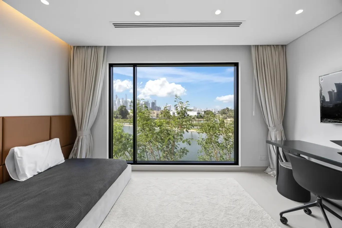 Modern bedroom with gray bedding, a tan headboard, and a large window overlooking a river and city skyline.