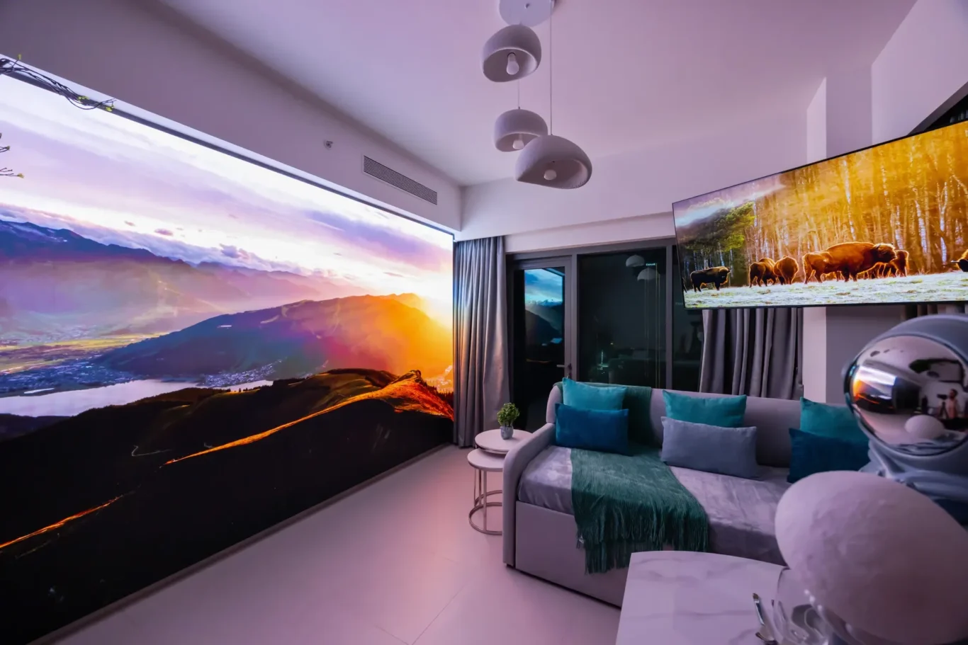 Living room with a panoramic mountain sunset mural, grey sofa with teal cushions, and a wall-mounted TV displaying grazing bison.