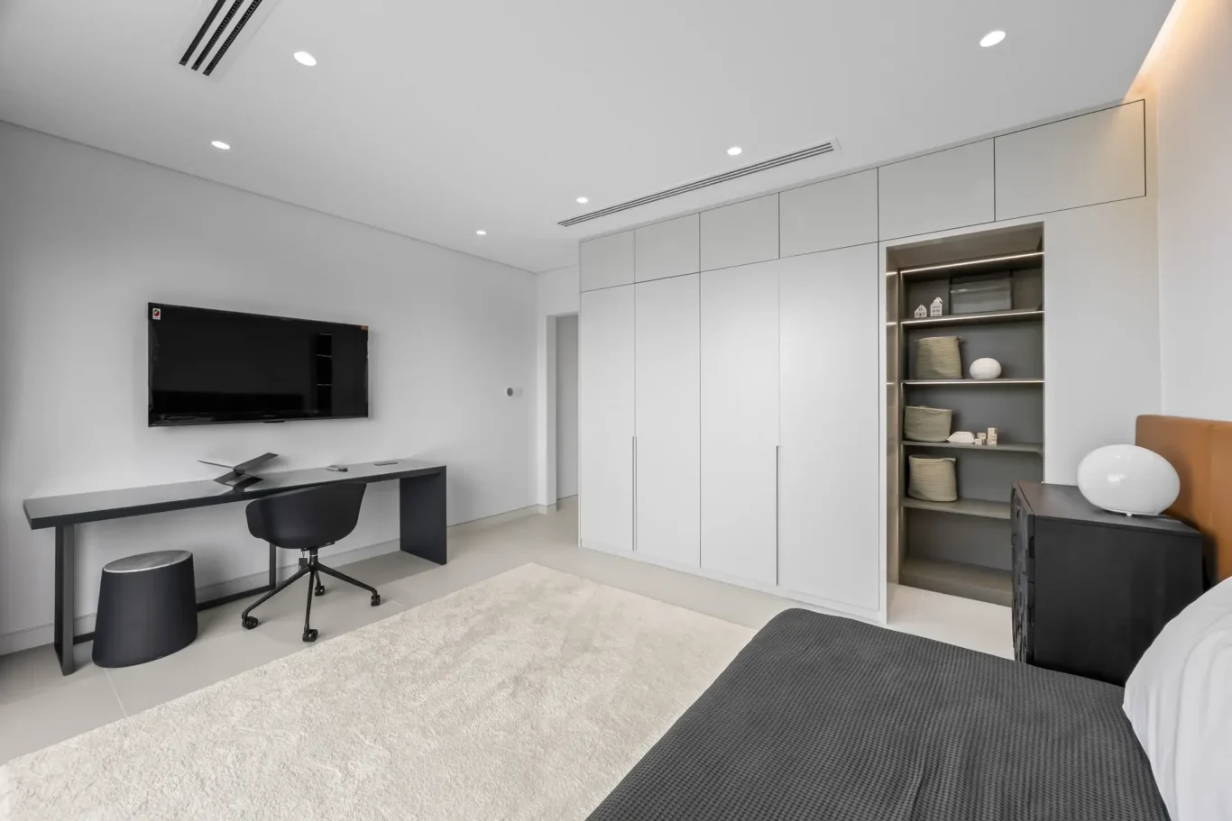 Modern bedroom with a wall-mounted TV above a black desk and chair, and a tall white built-in wardrobe with doors on the right.