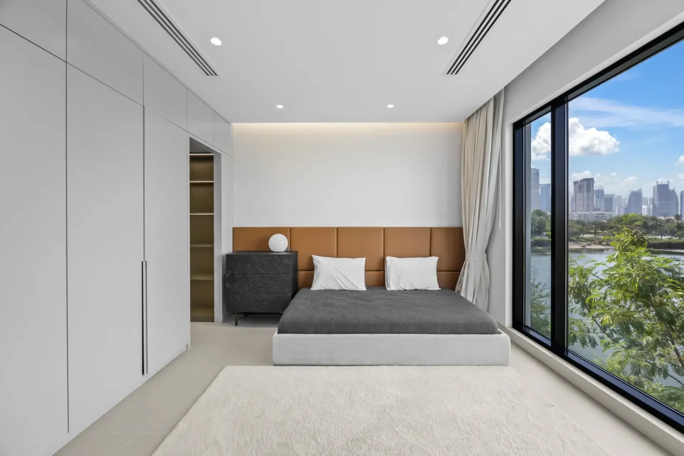 Modern minimalist bedroom with built-in white wardrobes on the left, a low platform bed with a tan headboard, and large windows showing a city skyline and river view.