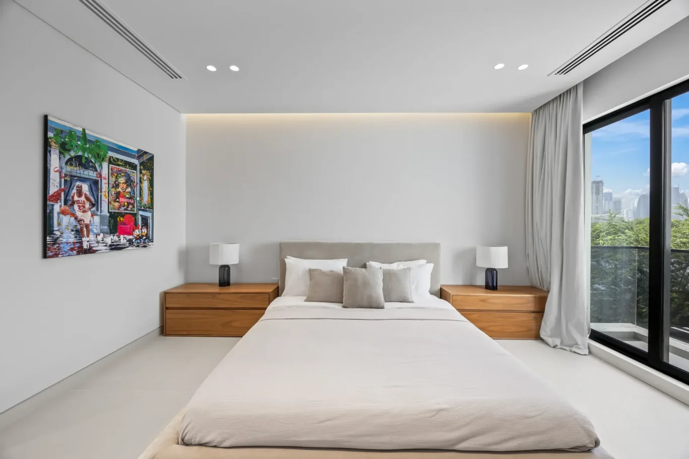 Bed centered in a bright modern bedroom with neutral bedding and two wooden nightstands, each with a white lamp, and a colorful framed artwork on the left wall.