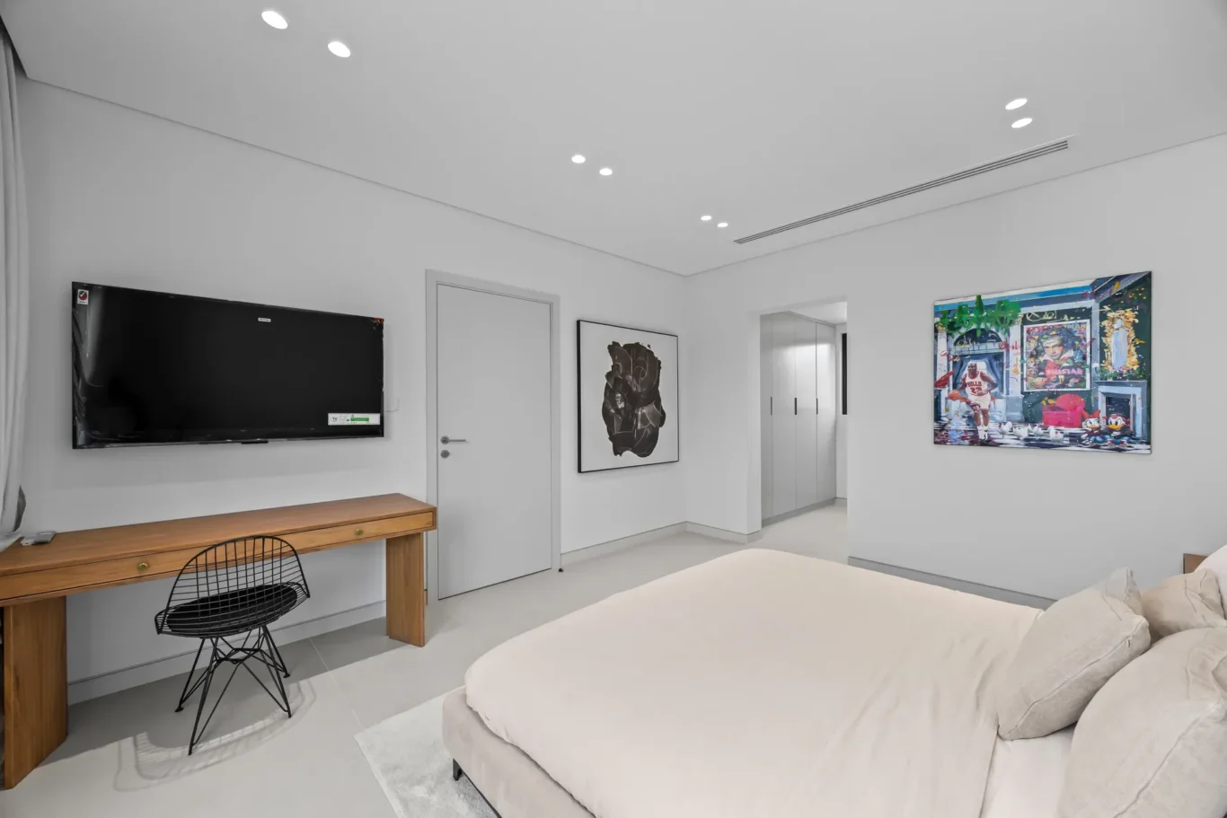 Modern bedroom with a wall-mounted TV, wooden desk, and black metal chair, white walls, and a large colorful artwork on the right.