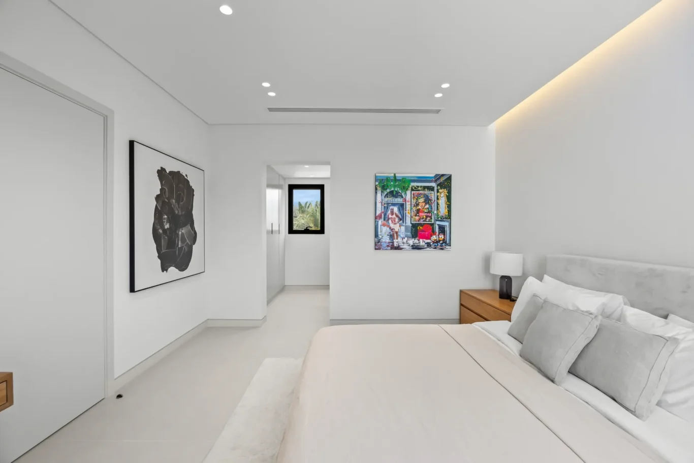 Bright minimalist bedroom with white bedding, gray pillows, and wall art; a hallway with a window is visible in the background.