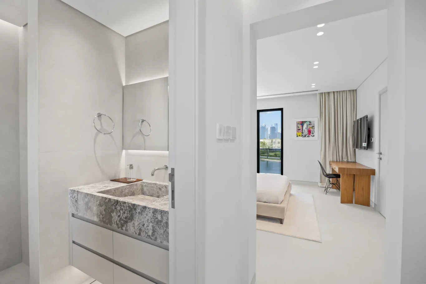 Open doorway into a bright, modern apartment showing a marble bathroom vanity on the left and a living area with sofa, desk, and TV on the right.