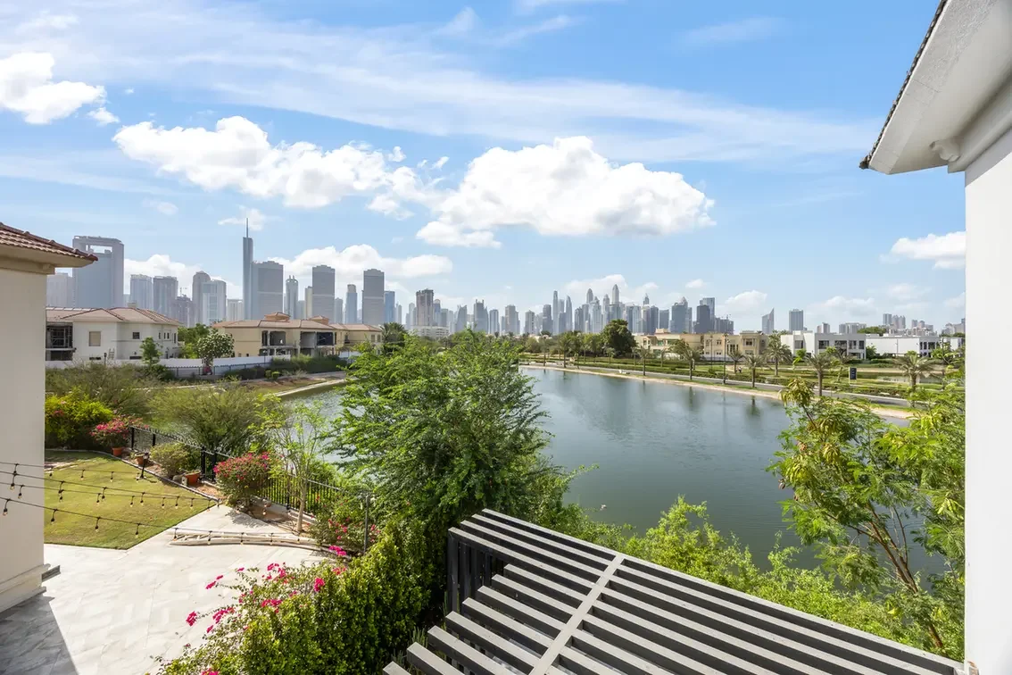 Balcony view over a canal toward a distant city skyline, with green lawns and houses in the foreground.