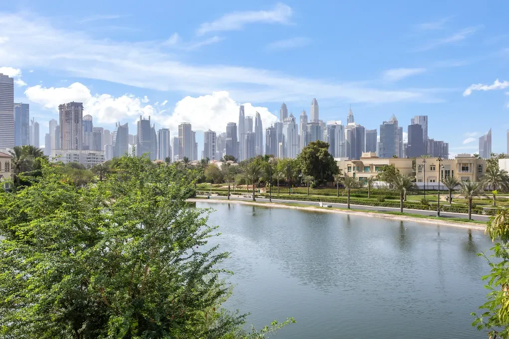City skyline with tall modern skyscrapers across a calm lake, with a tree-filled foreground and a green park along the water edge.