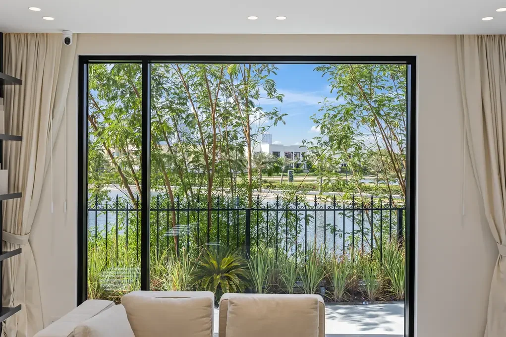Living room with a large glass sliding door framing a garden and water beyond, beige seating in the foreground.