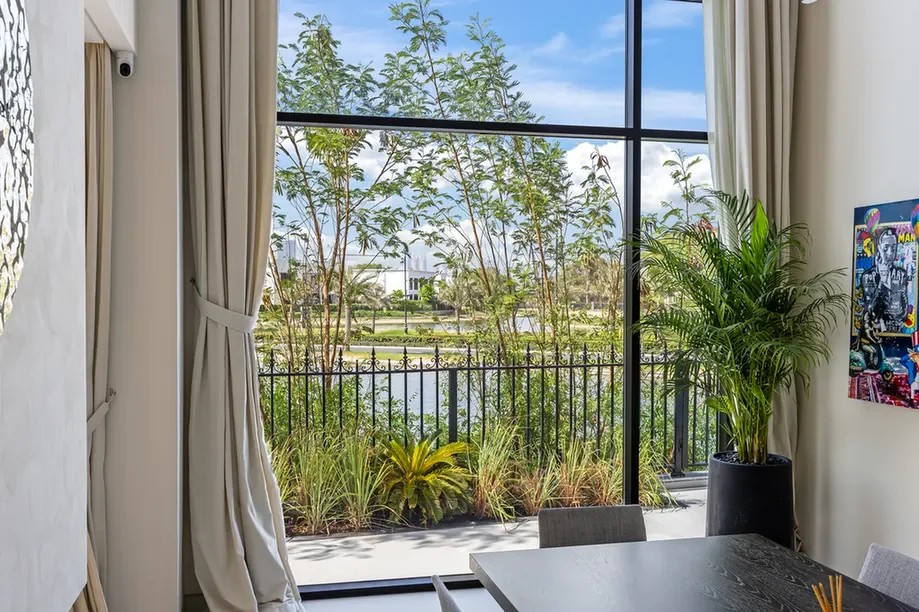 Bright dining area with a large floor-to-ceiling window overlooking a garden and pond, with potted plants and beige curtains nearby.