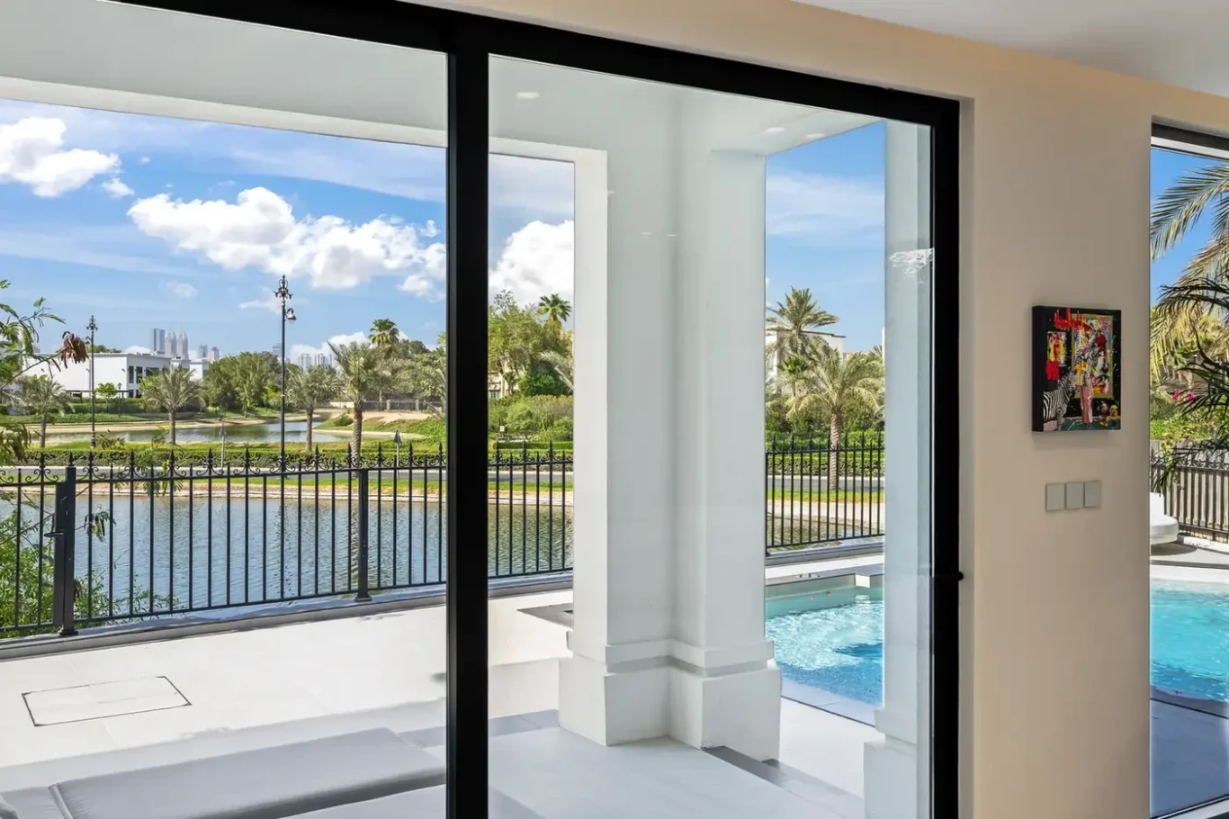 Interior view through large glass doors opening to a pool area, with a lake, palm trees, and railing beyond.