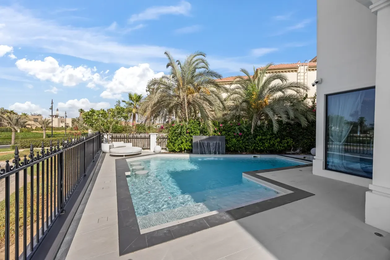 Modern backyard pool area with palm trees, loungers, and a black metal fence along a sunny patio edge.