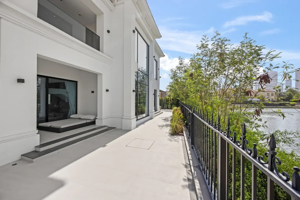 Modern white building with a patio and outdoor seating by a glass door, overlooking a railing and river with trees.