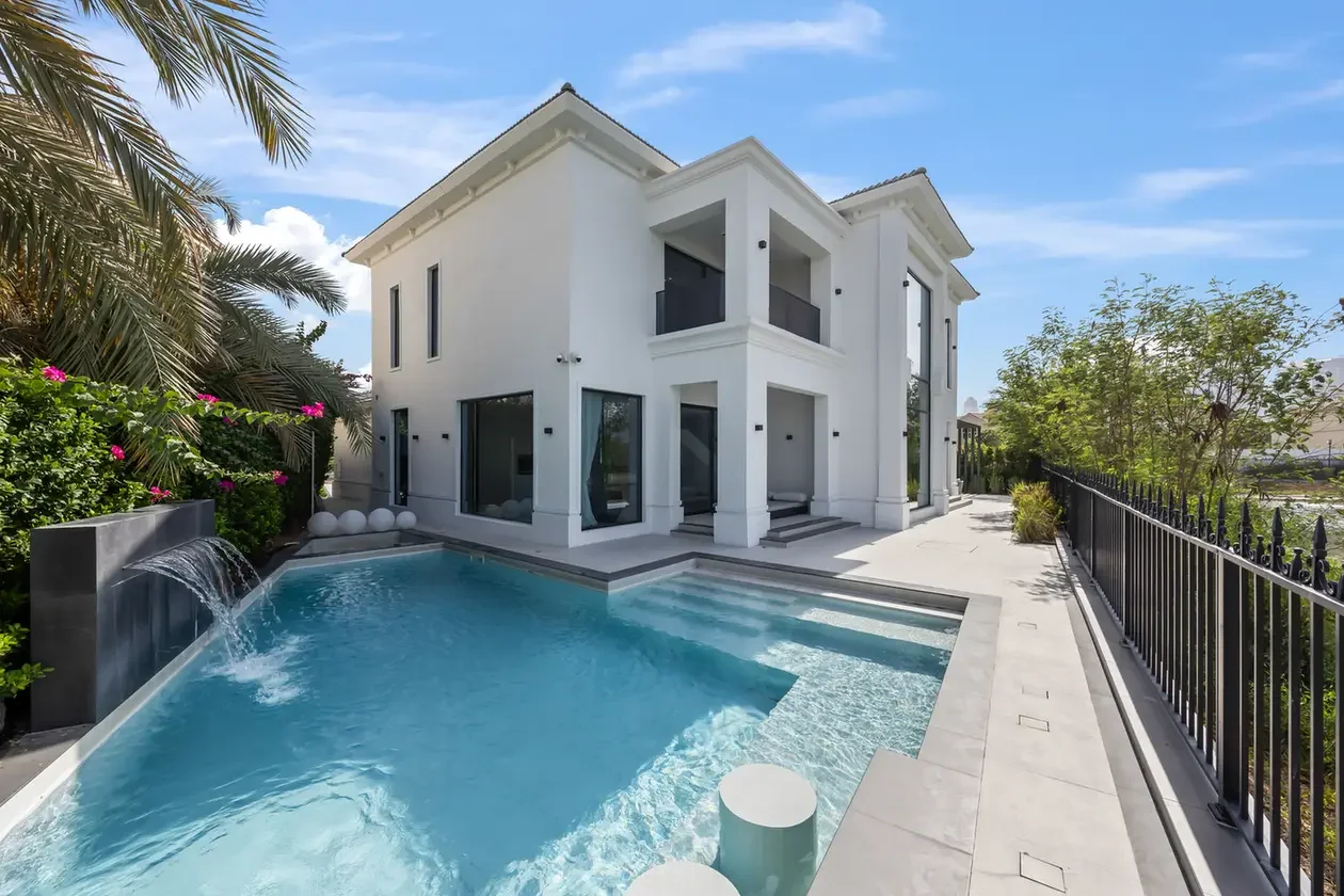 Modern white two‑story villa with a rectangular pool, small fountain, and palm trees on the left under a blue sky.
