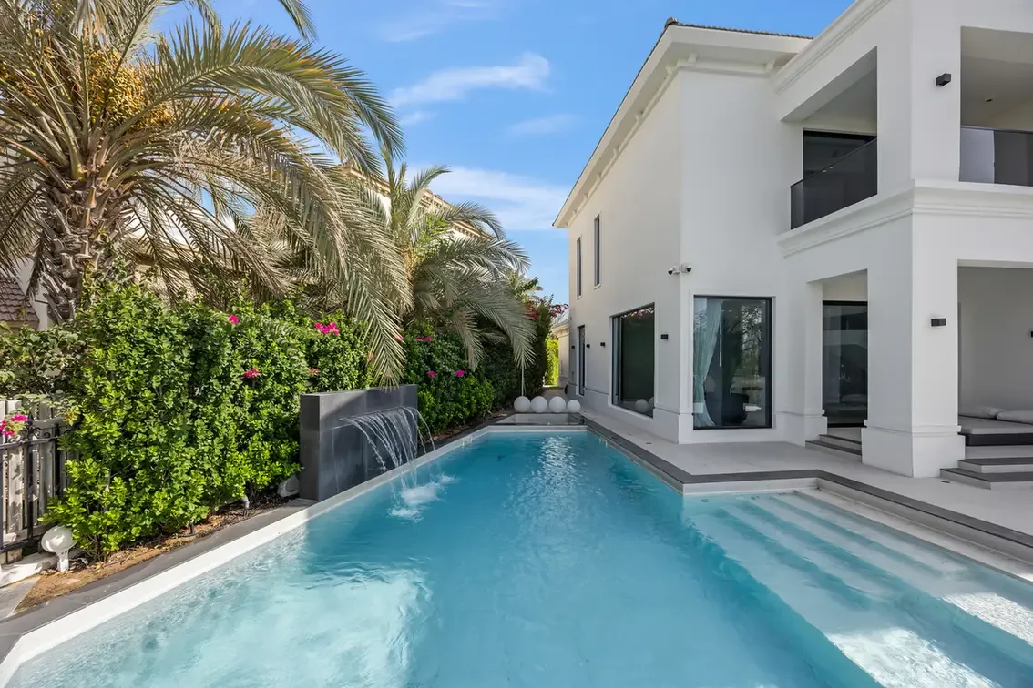 Modern white two-story house with large glass doors beside a rectangular swimming pool, palm trees and greenery along the fence.