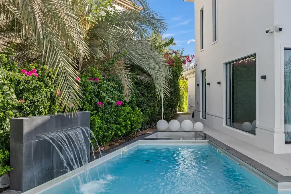 Modern backyard with a rectangular pool, a waterfall feature, palm trees, and a white contemporary house nearby.