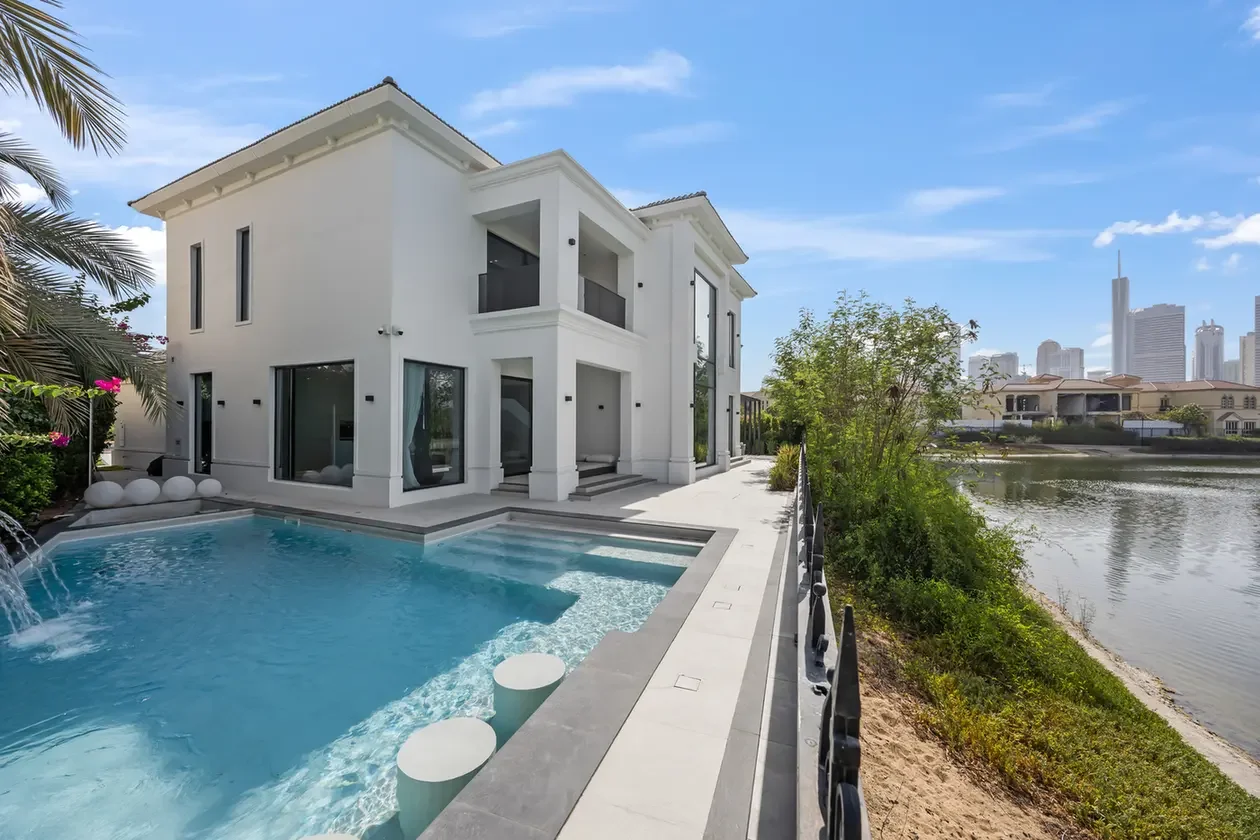Modern white two-story villa with a rectangular pool, terrace, and water feature by a river; high-rise city skyline in the distance.
