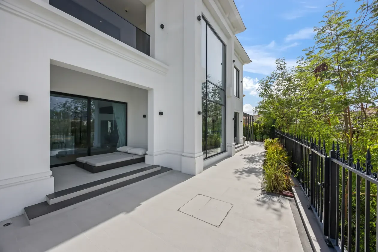 White modern house terrace with large glass doors, outdoor bed on steps, and a black metal fence beside potted plants.