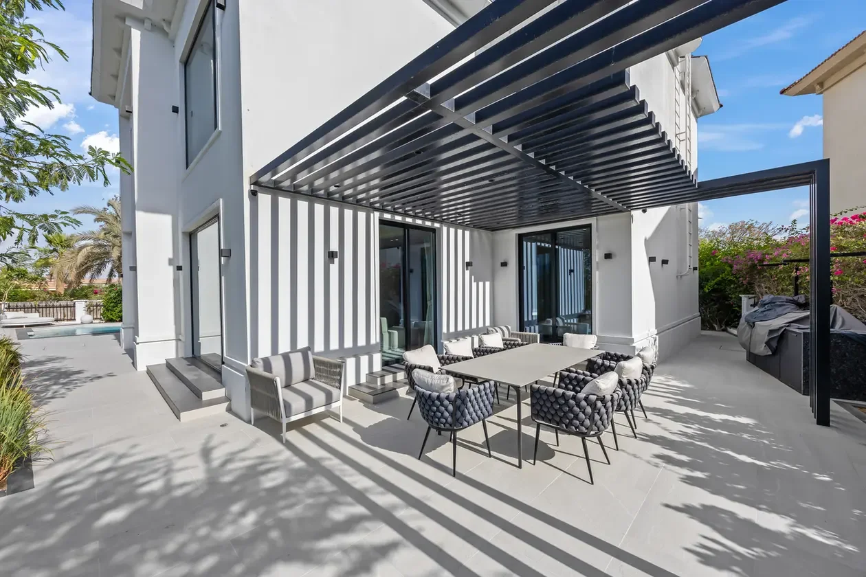 Modern white house back patio with a large rectangular dining table surrounded by eight cushioned wicker chairs under a black slatted pergola, sunny outdoor setting.