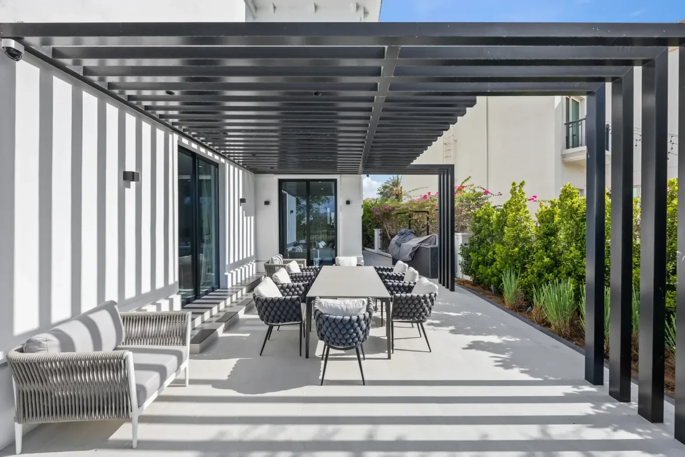 Outdoor patio with a long dining table and eight woven chairs under a black pergola beside a white house.