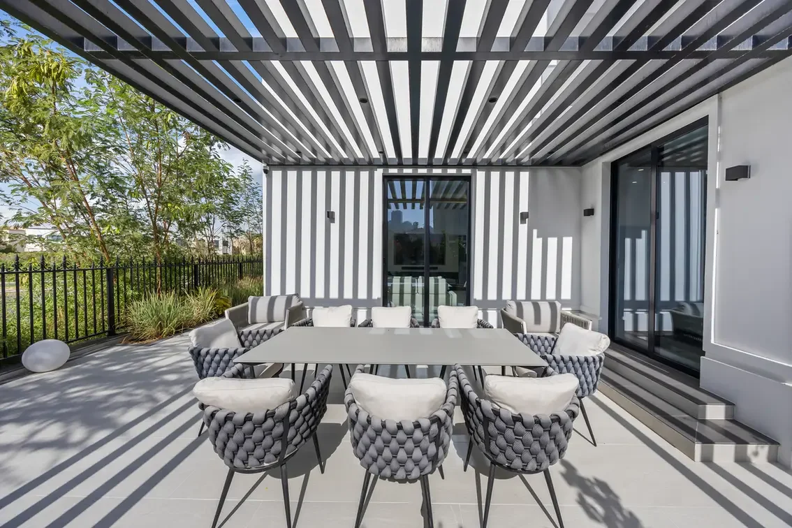 Outdoor dining patio under a slatted pergola, eight gray woven chairs around a rectangular table, with glass doors and greenery outside.