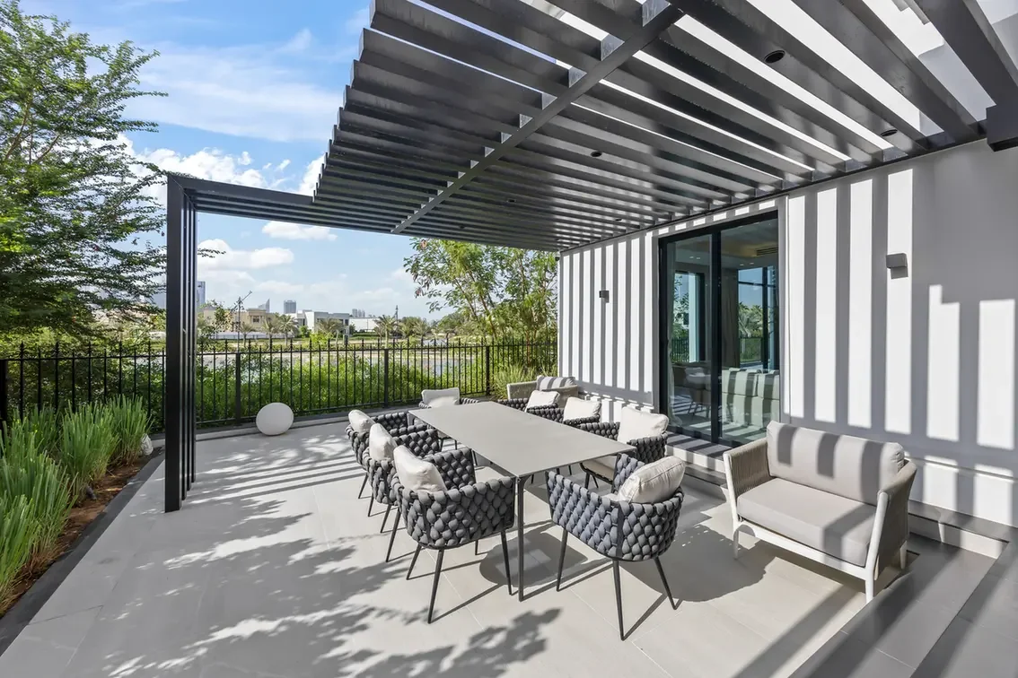 Modern outdoor patio with a long rectangular dining table and cushioned chairs under a slatted pergola on a balcony overlooking greenery and a distant city skyline.