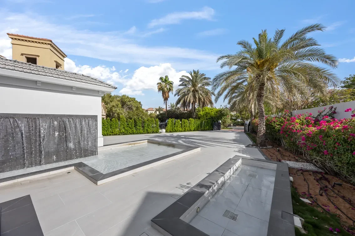 Modern courtyard with a cascading water wall, shallow pool, palm trees, and pink flowering bushes under a blue sky.