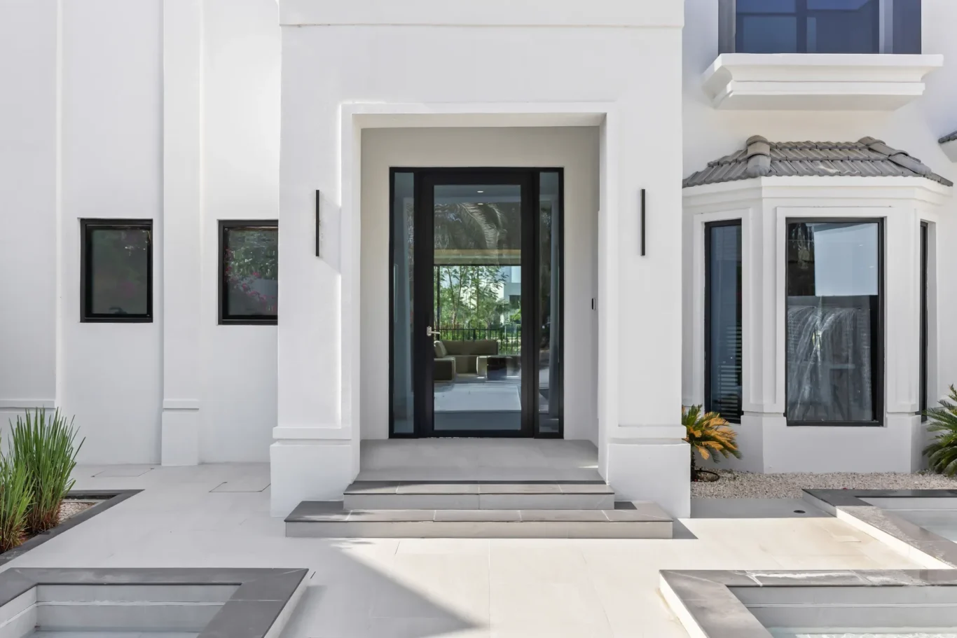 White contemporary house entrance featuring a black-framed glass door, tall side columns, and a reflecting courtyard with patios and planters