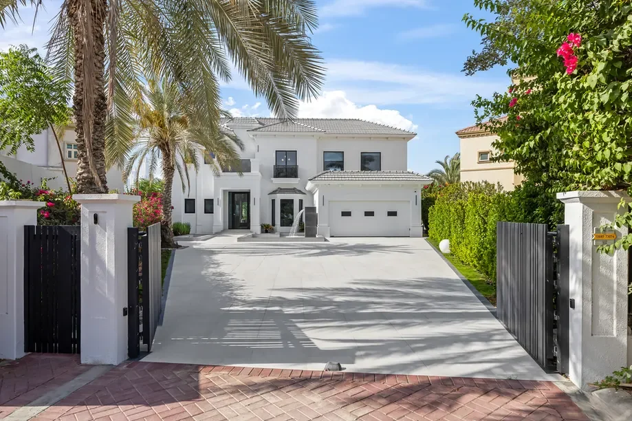 White two-story house with a wide concrete driveway and a double garage, gated entry, and palm trees in a sunny yard.