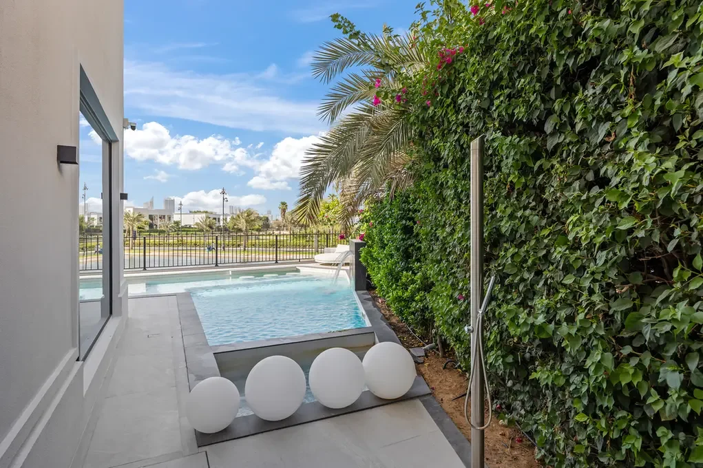 Modern poolside patio with a rectangular pool, white spherical decor on the deck, an outdoor shower, and a tall green hedge nearby.