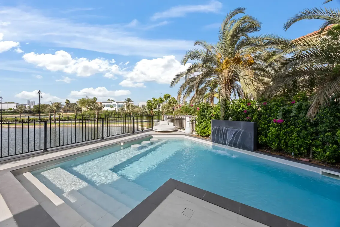 Luxurious backyard pool with steps, a stone deck, and a cascading water feature, bordered by palm trees and a black fence overlooking a canal.