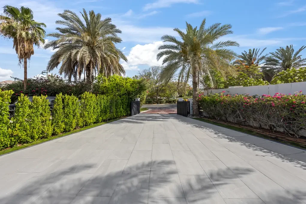 Wide driveway lined with green hedges and palm trees leading to a gated entrance under a blue sky.