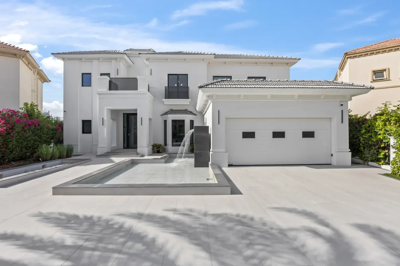 Front view of a modern white two-story villa with a water feature and a two-car garage against a blue sky.