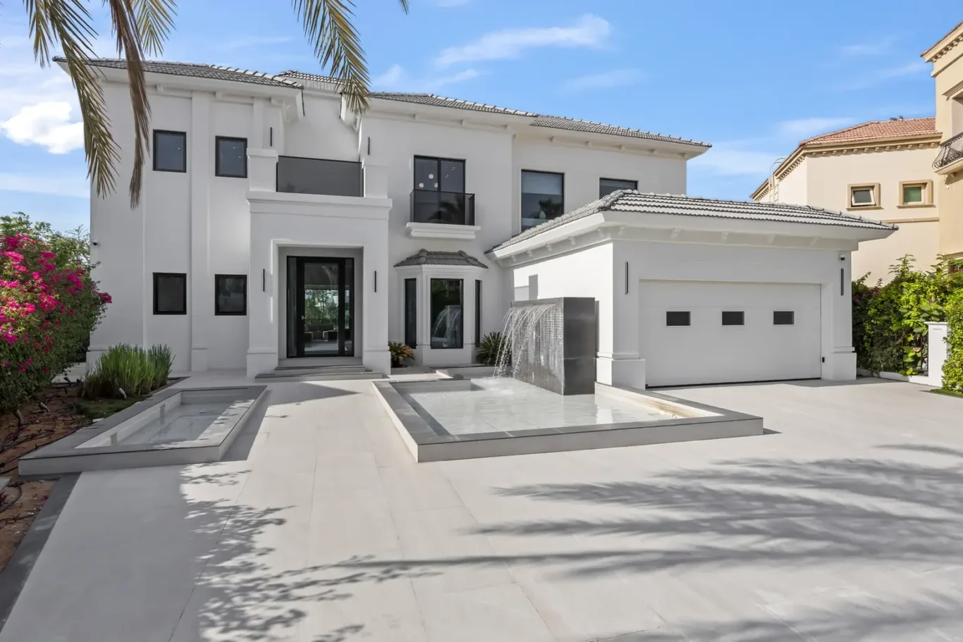 White contemporary two‑story house with a front water feature, reflecting pools, and a two‑car garage, framed by palm trees.