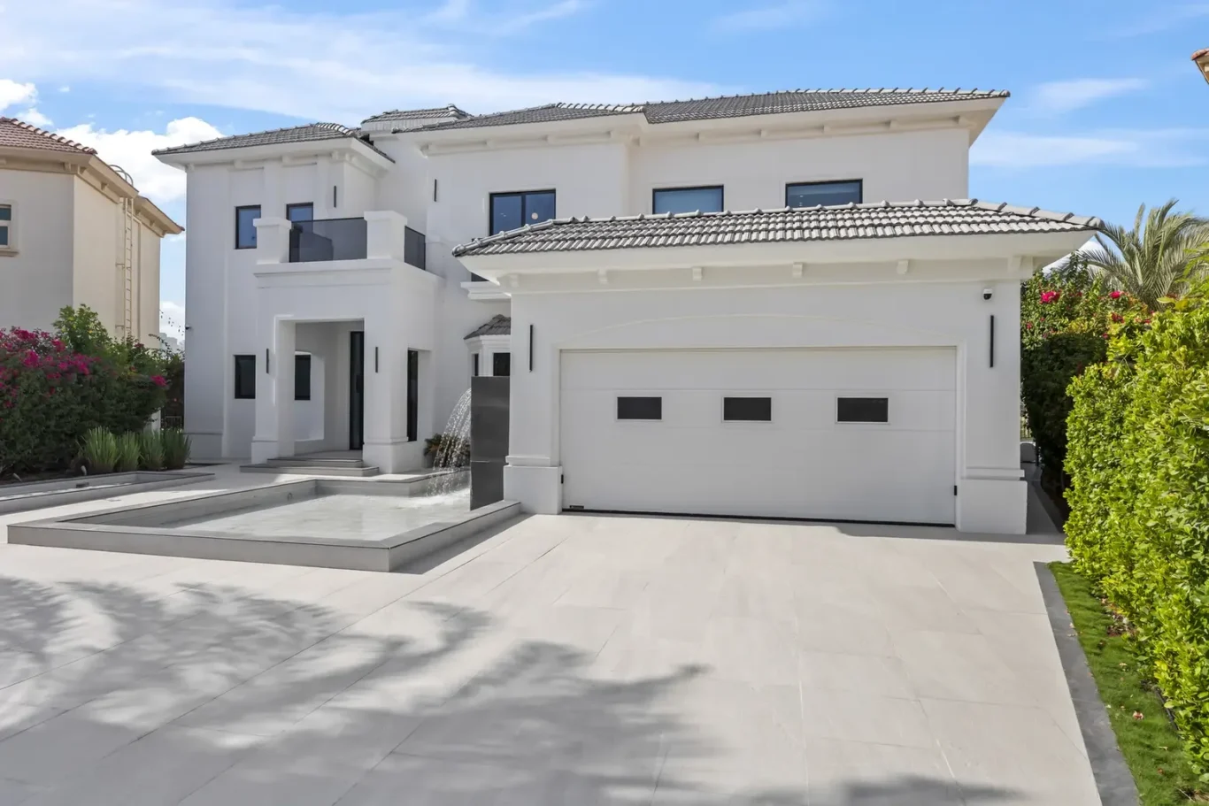 White two-story house with a two-car garage, a rectangular water feature in the front yard, and trimmed hedges under a blue sky.