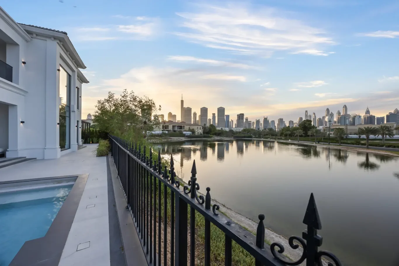 White modern house with glass doors beside a narrow pool, looking toward a calm river and a distant city skyline at sunset.