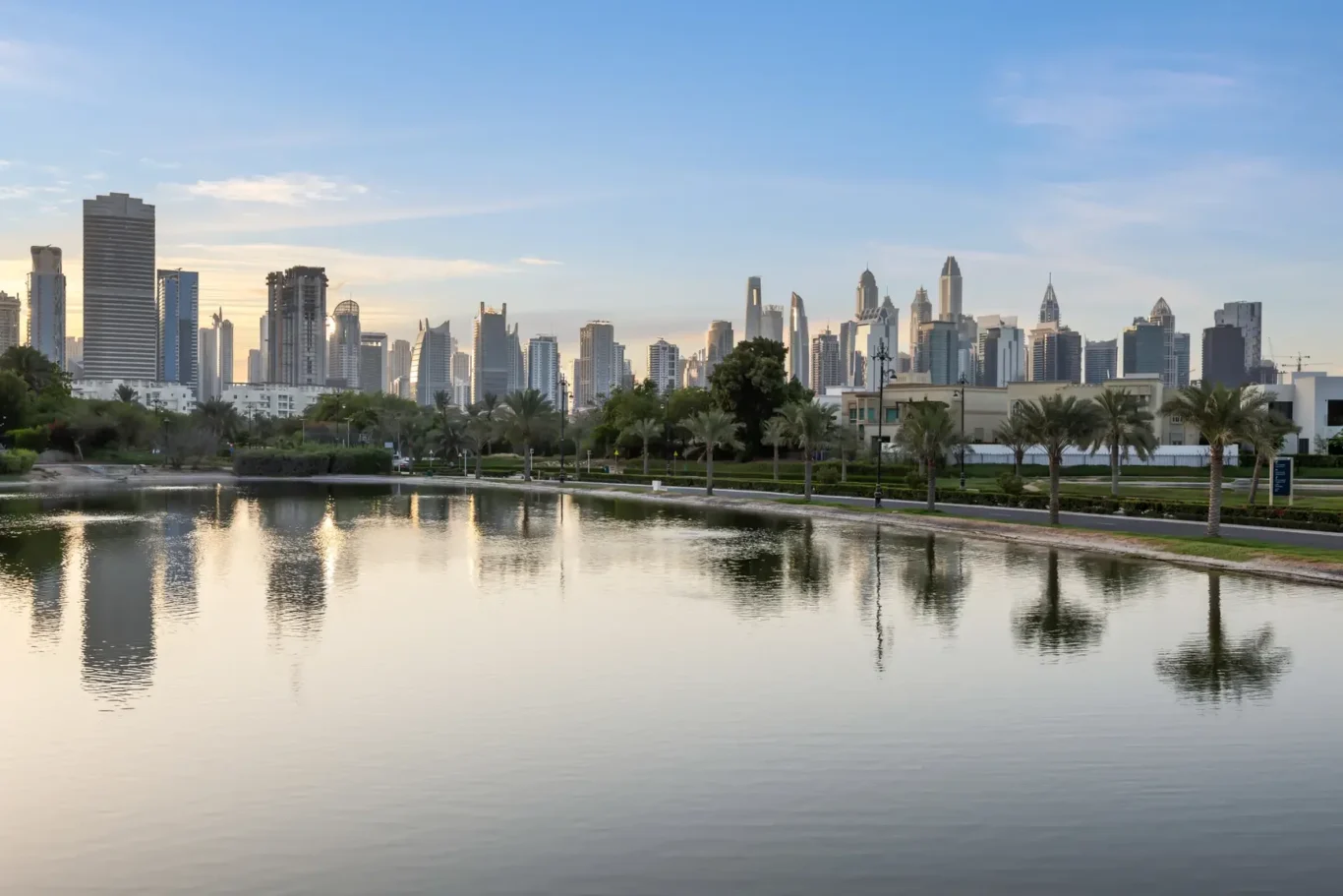 Dubai skyline with tall glass buildings along a calm waterfront park at dusk, palm trees lining the shore and their reflections in the water