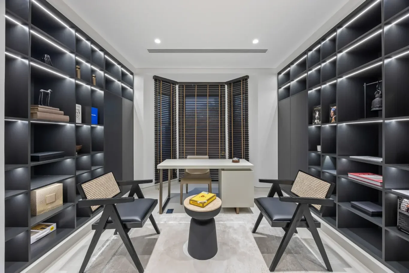 Modern home office with dark wall-to-wall shelves, a white desk centered between two black chairs, and a window with wooden blinds.