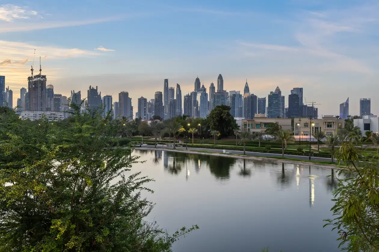 City skyline of tall modern buildings across a calm river at dusk, with green trees and a park along the water’s edge in the foreground.