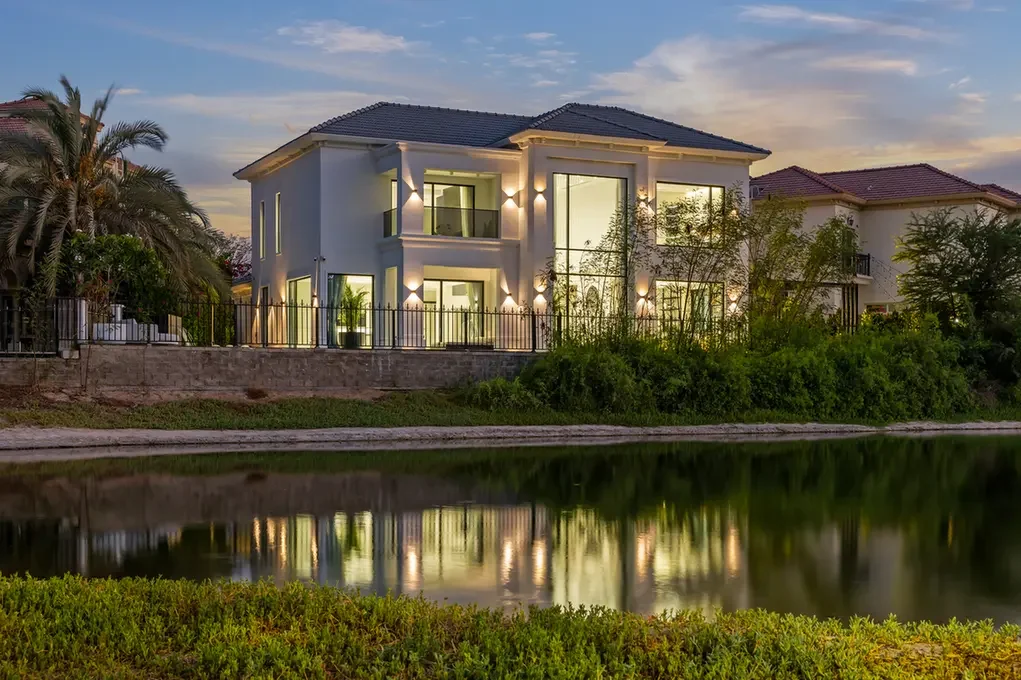 Modern two-story house with warm exterior lights along a fence, by a calm canal with reflection and landscaping at sunset/dusk