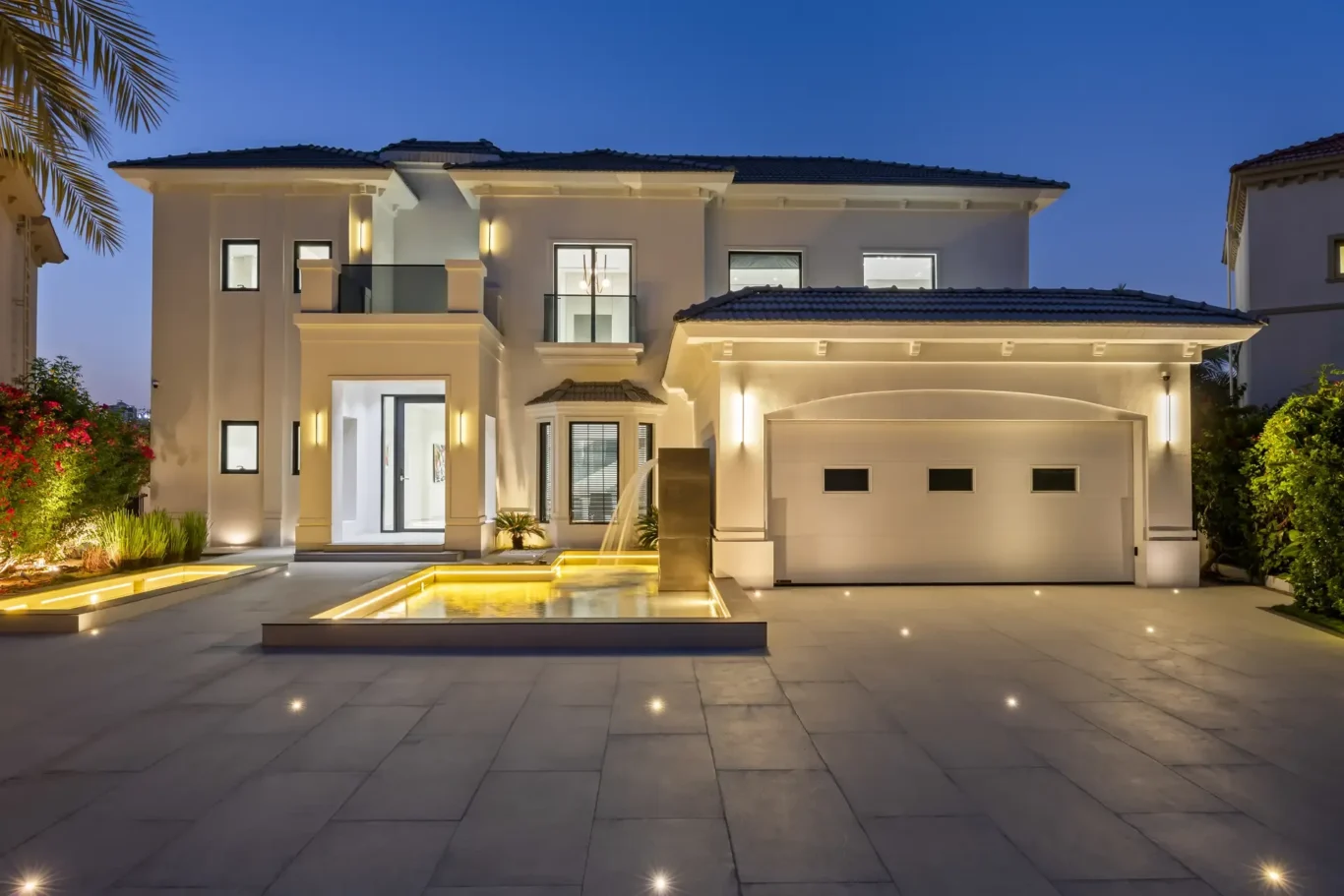 Modern two-story white house at dusk with illuminated entry and palm-fringed landscaping on the left.