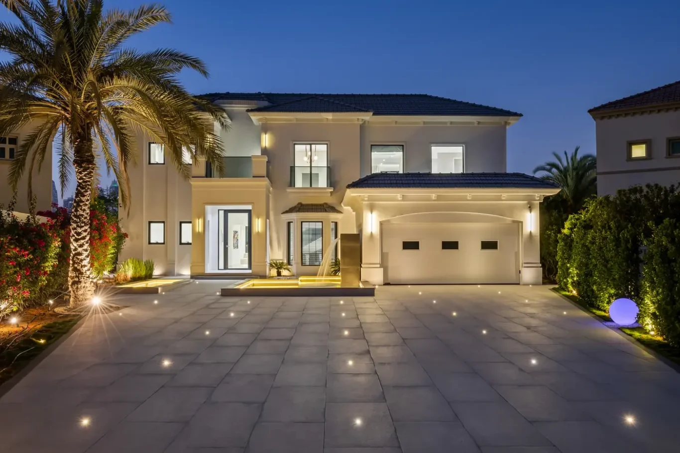 Night view of a two-story modern villa with a lit entrance, wide tiled driveway, and a two-car garage.