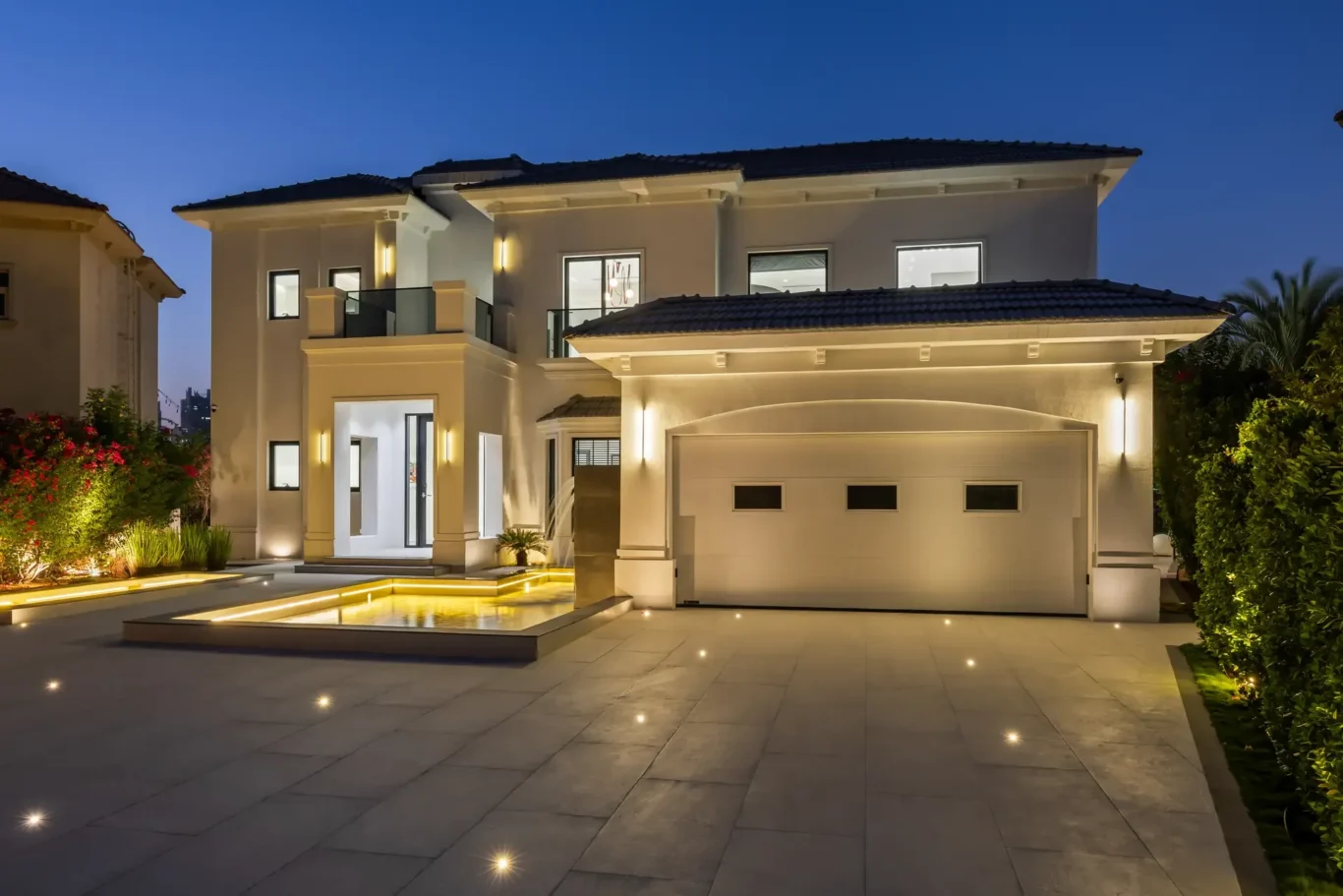 Modern two-story house at night with warm exterior lighting and a lit courtyard, driveway, and landscaped garden surrounding a reflecting pool area