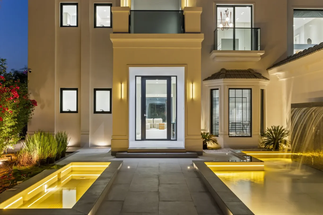 Modern two-story beige villa at night with a lit entryway, glass door, and luminous rectangular water features along a paved path leading to the entrance still reflecting warm lighting.