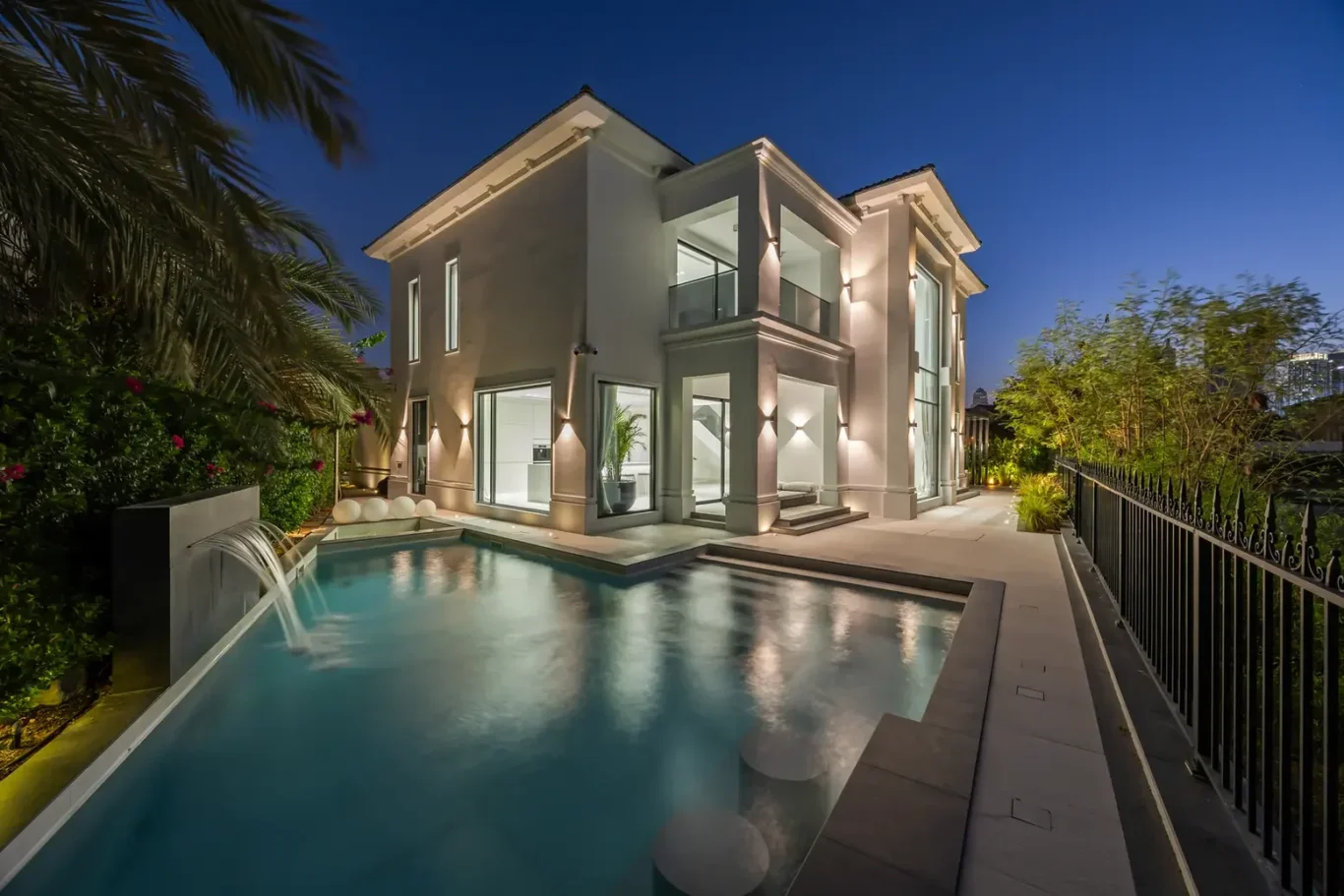 Modern two-story white villa with glass walls and balconies, illuminated at night beside a rectangular pool.