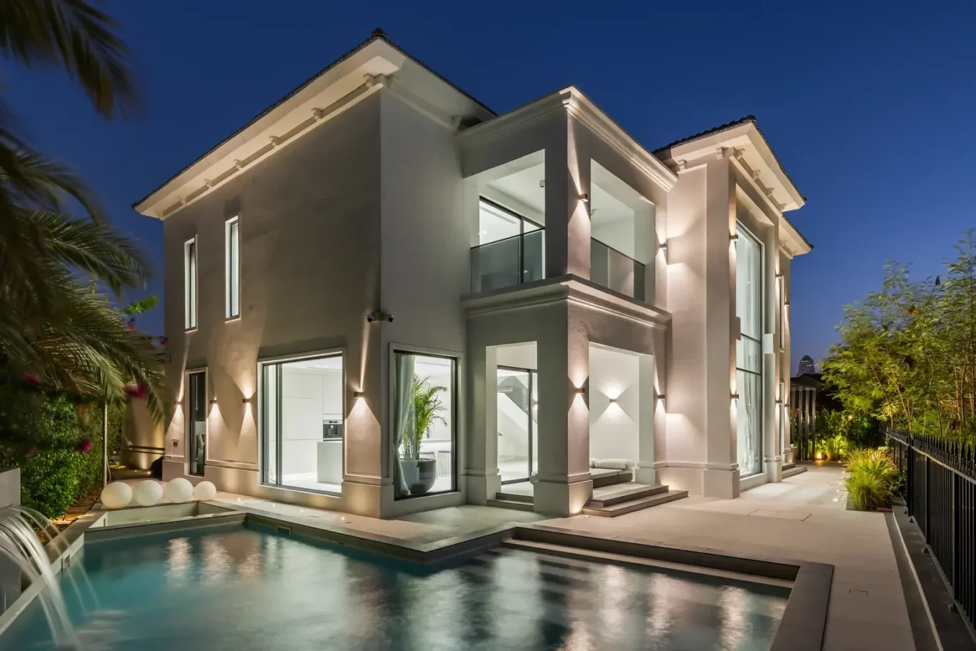 Modern two‑story house with white exterior lights, glass walls and a rectangular pool at dusk. nearby tropical plants frame the scene.